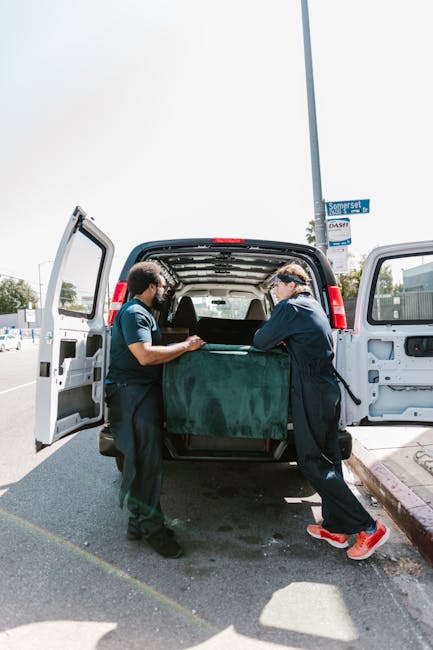 Two movers from Man With a Van Aldgate are engaged in a home relocation process, loading a large piece of furniture covered with a green protective blanket into the back of a white van parked on a res