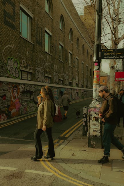 A busy street scene outside a building with brickwork and graffiti murals, where pedestrians are crossing the road and walking along the pavement. In the foreground, a woman with curly hair, wearing a green jacket and black trousers, is standing near the curb, holding a mobile phone to her ear. Adjacent to her, a man in a dark jacket, baseball cap, and carrying a backpack is walking past a lamppost covered with posters and stickers, with a small dog on a leash nearby. In the background, other people are visible walking along the sidewalk, some heading towards the street, with the environment lit by natural daylight. The setting appears to be part of a busy urban area, consistent with the location described in the Removals in Aldgate East and Spitalfields Market page, reflecting typical street activity in this district managed by Man With a Van Aldgate during home relocation or moving logistics.