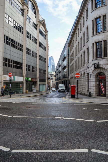 A narrow city street in Aldgate, London, with tall stone and glass office buildings lining both sides. The road features painted white lines and zebra crossings, with a few vehicles including a white van parked or moving along the street. A red public telephone booth and a collection of blue and red signage are visible on the right side, with pedestrians present on the left sidewalk. The sky above is partly cloudy, illuminating the urban environment. The scene is associated with the context of home relocation and furniture transport, capturing the typical street environment where professional removals, such as those provided by Man With a Van Aldgate, operate during packing and moving processes.